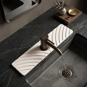 May include: A rectangular, off-white silicone faucet splash guard with diagonal ridges positioned behind a dark bronze metal faucet. The faucet is mounted on a dark stone countertop above a dark gray sink basin with a metal drain grate.