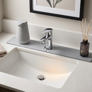 May include: A rectangular gray silicone faucet splash guard mat positioned behind a chrome bathroom faucet. The mat features a central cutout for the faucet base and provides a flat surface for a gray soap dispenser and a glass reed diffuser.