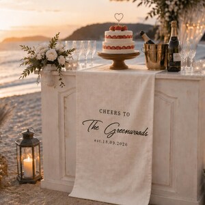 Puede incluir: Una mesa de bar de madera blanca cubierta con un camino de mesa de tela color crema con el texto impreso en negro "CHEERS TO The Greenwoods est. 18.09.2026". La mesa sostiene un pastel de fresas de dos niveles, una cubitera dorada y copas.