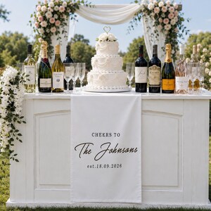 Puede incluir: Una mesa de bar de madera blanca con un camino de mesa colgante que muestra el texto negro "CHEERS TO The Johnsons est.18.09.2026". Sobre la mesa hay un pastel de bodas blanco de tres niveles, botellas de vino y copas de cristal.