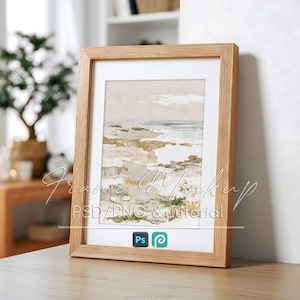 May include: A light wood picture frame containing an abstract landscape print with beige, tan, and muted green tones. The frame features a white mat board. Text overlay reads Frame Mockup PSD/PNG & Tutorial with Adobe Photoshop and Procreate icons.