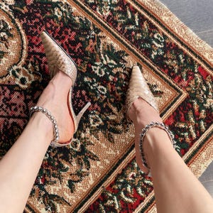 Könnte beinhalten: Ein Paar beigefarbene Pumps mit spitzer Zehenpartie, Stiletto-Absatz und einer strukturierten Schlangenleder-Optik. Jeder Schuh verfügt über einen zarten Knöchelriemen, der mit einer Reihe klarer, runder und facettierter Kristalle besetzt ist.