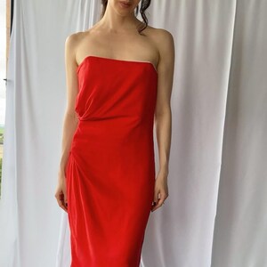 May include: A vibrant red strapless midi dress featuring a straight neckline and subtle side ruching detail. The garment is constructed from a smooth, solid-colored fabric that falls to a mid-calf length, creating a clean and streamlined silhouette.