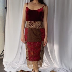 Può includere: Un abito midi con corpetto in velluto rosso scuro a spalline sottili, un'ampia fascia in pizzo dorato in vita e una gonna patchwork che combina tessuto marrone testurizzato, broccato floreale rosso e un orlo in pizzo dorato.