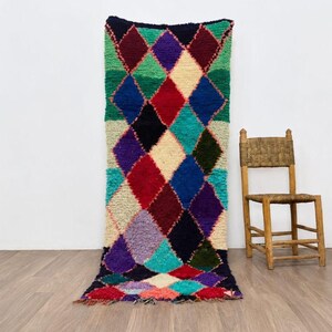 Peut inclure: Un tapis rectangulaire en laine présentant un motif en losanges multicolore et vibrant. Le design se compose de formes imbriquées dans des tons de rouge, bleu, vert, violet, noir et crème. Le tapis a un poil épais et texturé avec des franges.