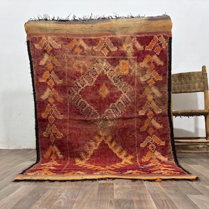 May include: A rectangular area rug featuring a deep red base color with intricate geometric patterns in orange, yellow, and dark brown. The rug has a thick, textured pile, a wide horizontal band at the top, and dark fringe along the edges.