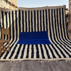 Peut inclure: Un grand tapis rectangulaire présentant un motif de rayures verticales noires et blanches avec un bloc rectangulaire bleu uni au centre. Le tapis possède des franges sur les bords supérieur et inférieur et est présenté avec une petite chaise en bois.