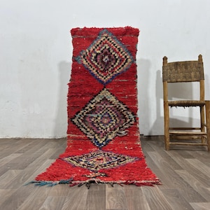 May include: A rectangular runner rug featuring a vibrant red base color with three large, centered diamond-shaped patterns. Each diamond contains intricate, multi-colored geometric details in shades of blue, yellow, black, and white, with a shaggy texture.