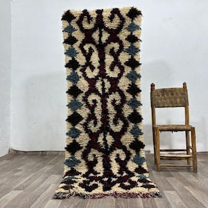 May include: A rectangular wool rug featuring a cream-colored base with a central vertical pattern of dark maroon, interconnected scroll-like shapes. The borders are decorated with a repeating diamond motif in alternating dark brown and blue-gray.