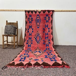 May include: A rectangular area rug featuring a vibrant red base with intricate geometric patterns in blue and dark brown. The design includes diamond shapes and triangular borders, finished with a fringed edge at the bottom and a textured pile.