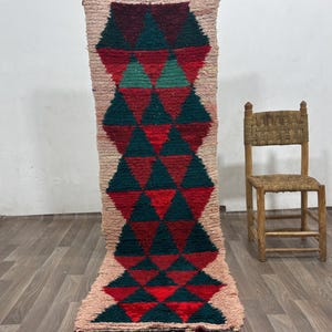 Peut inclure: Un long tapis rectangulaire orné d'un motif géométrique répétitif composé de triangles vert foncé et rouge profond. Le tapis présente une texture à poils longs et épais avec des bordures beige clair et une extrémité frangée.
