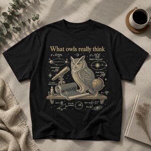 May include: A black t-shirt featuring a gold-toned graphic of an owl perched on a shelf with a telescope, a globe, and a scroll. The text "What owls really think" is printed above, surrounded by various physics and astronomy equations.