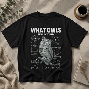 May include: A black short-sleeve t-shirt featuring a white graphic of an owl perched on a branch. The text reads WHAT OWLS REALLY THINK, surrounded by scientific diagrams, mathematical formulas, and the names Johannes Kepler, Isaac Newton, and Edwin Hubble.