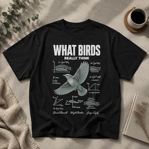 May include: A black short-sleeve t-shirt featuring a white graphic print with the text WHAT BIRDS REALLY THINK. The design includes a central bird illustration surrounded by various physics equations, aerodynamic diagrams, and labels like Daniel Bernoulli.
