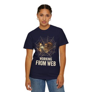 May include: A navy blue t-shirt featuring a graphic of a brown spider sitting in a web while using a laptop under a small lamp. The text "WORKING FROM WEB" is printed in large, cream-colored capital letters below the spider illustration.