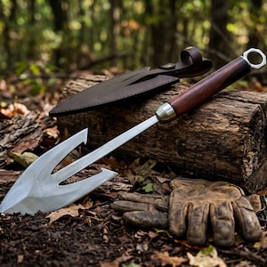 May include: A metal garden trowel with a three-pronged fork head and a dark wood handle featuring a silver hexagonal nut at the end. The tool includes a dark brown leather sheath with a strap and a pair of worn brown leather work gloves.