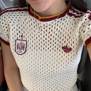 May include: A cream-colored crochet short-sleeve top featuring a maroon and yellow ribbed neckline and sleeve cuffs. The shirt displays a red and white embroidered Spanish national crest on the right chest and a red embroidered trefoil logo on the left.
