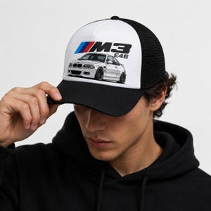 Peut inclure: Une casquette trucker noire et blanche avec une impression graphique d'une voiture BMW M3 E46 blanche sur le panneau avant. La casquette possède une visière noire unie, un panneau avant blanc avec le logo M3 E46 en noir, bleu et rouge, et des panneaux arrière en maille noire.