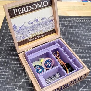 Puede incluir: Una caja de puros de madera Perdomo con la tapa abierta que muestra una ilustración de paisaje y el texto Perdomo Lot 23 Esteli, Nicaragua. El interior contiene un organizador de plástico morado claro con recipientes, alfileres y cables.