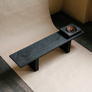 May include: A long, rectangular black wooden bench with a visible grain texture, supported by two thick vertical legs. On the right side of the bench sits a square black wooden block featuring a circular indentation containing a small brown bowl.
