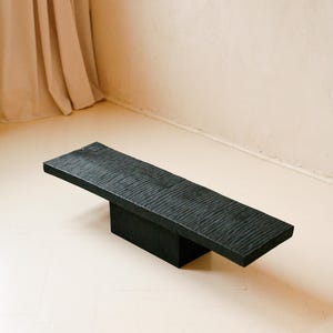 Puede incluir: Un banco bajo rectangular de color negro que presenta una superficie texturizada y ondulada en el panel superior. La pieza se apoya sobre una base central única, sólida y en forma de bloque, creando una estructura horizontal equilibrada.