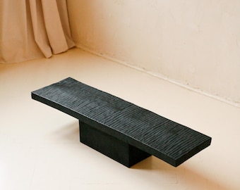 May include: A low, rectangular black bench featuring a textured, rippled surface on the top panel. The piece is supported by a single, solid, block-like central base, creating a balanced, horizontal structure with a dark, matte finish throughout.