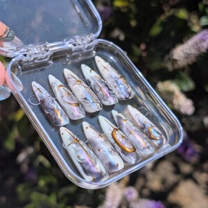 Puede incluir: Un juego de diez uñas postizas en forma de estilete dentro de un estuche de plástico transparente. Las uñas presentan una base marmolada en blanco, gris y morado con detalles celestiales en oro metálico, estrellas, lunas y puntos.