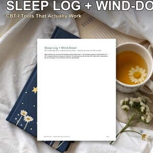 May include: A white document titled Sleep Log + Wind-Down with CBT-I text, placed on a dark blue notebook with gold star and flower illustrations. Beside it sits a white mug filled with tea and a small sprig of white flowers on white fabric.