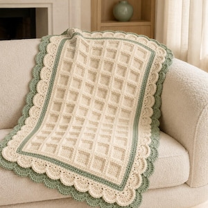 May include: A rectangular crochet blanket featuring a cream-colored waffle stitch pattern in the center. The blanket is bordered by a thin sage green stripe and finished with a wide, scalloped edge in alternating cream and sage green yarn.