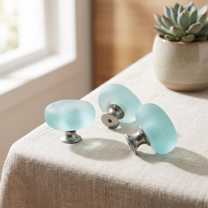 May include: Three cabinet knobs featuring smooth, oval-shaped, frosted light blue glass heads attached to brushed silver-toned metal bases. The knobs have a rounded, translucent appearance and are designed for attachment to furniture or cabinetry doors.