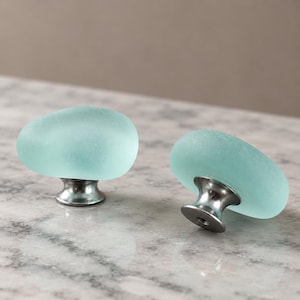May include: Two cabinet knobs featuring smooth, frosted, seafoam green glass tops shaped like rounded pebbles. Each knob is attached to a polished silver-toned metal base with a circular mounting point, designed for installation on furniture or cabinetry.