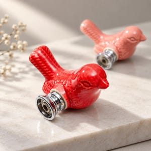 Puede incluir: Dos pomos para muebles con forma de pájaro, uno en rojo vibrante y otro en rosa coral suave. Cada pájaro de cerámica presenta un diseño detallado y texturizado en alas y cola, montado sobre una base de metal plateado pulido con un orificio.