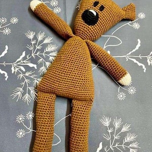 May include: A brown crocheted stuffed animal with a triangular head, pointed ears, and a black circular nose. The toy features long limbs with white tips on the hands and feet, and a textured yarn construction throughout its entire body.