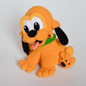 Op de afbeelding: Een gehaakt knuffeldier in de vorm van een cartoonhond van oranje garen, met grote witte oogvlekken, zwarte oren, een zwarte neus, een groene halsband en een klein roze tongetje dat uitsteekt. Het figuurtje zit met de voorpoten naar voren.