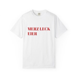 May include: A white short-sleeved t-shirt featuring the red text MERZ LECK EIER printed in a bold serif font across the center of the chest. The garment has a classic crew neckline and a standard fit, showcasing a clean and simple design.