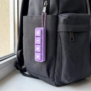 Puede incluir: Un llavero rectangular de color morado claro con cuatro teclas cuadradas dispuestas verticalmente, cada una con una letra blanca que forma la palabra KIRA. El llavero está sujeto a la cremallera de una mochila gris oscuro con una cadena.