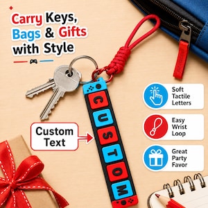 Puede incluir: Un llavero rectangular con bloques alternos rojos y azules con letras negras que forman la palabra CUSTOM, unido a una correa de muñeca trenzada roja y una anilla metálica con dos llaves. Texto: Carry Keys, Bags & Gifts with Style.