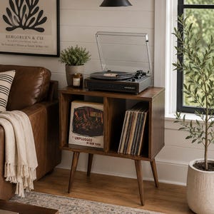 May include: A dark wood record player stand with four tapered legs and two open storage compartments. The top surface holds a black Crosley turntable with a clear dust cover. The shelves contain a Nirvana Unplugged in New York vinyl record.