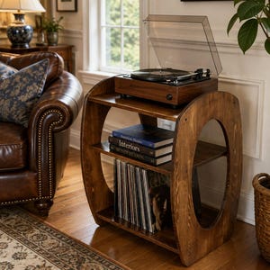 Puede incluir: Un soporte para tocadiscos de madera oscura con marco ovalado y tres niveles. La superficie superior sostiene un tocadiscos negro con cubierta transparente. El estante central contiene tres libros apilados y el inferior discos de vinilo.
