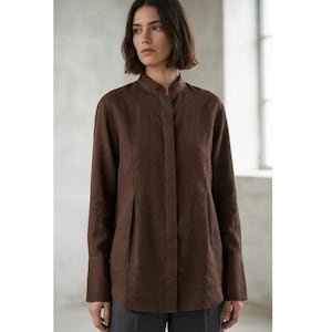 May include: A dark brown long-sleeved shirt featuring a mandarin collar, a concealed button placket, and vertical pleat detailing at the front. The garment has a straight hemline and wide cuffs, constructed from a textured fabric with a matte finish.