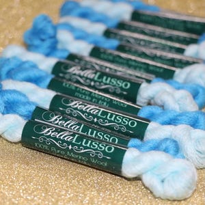May include: Close-up of several spools of blue and white ombre yarn with green labels that read "Bella Lusso 100% Pure Merino Wool made in Italy".