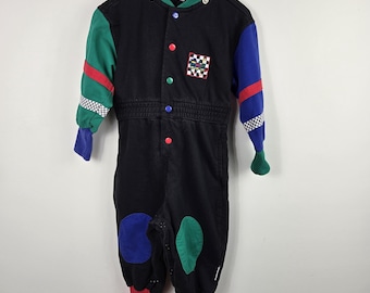 May include: A black long-sleeve jumpsuit featuring a green right sleeve, a blue left sleeve, and red cuffs. The front has a row of multicoloured snap buttons, a checkered patch, and a green collar. The legs include blue and green knee patches.