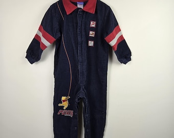 May include: A navy blue corduroy jumpsuit featuring a red collar, long sleeves with red and white stripes, and a front button closure. The garment displays a graphic of Winnie the Pooh, the text POOH, and three small square patches on the chest.