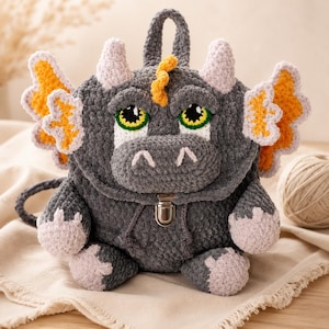May include: A gray crocheted backpack shaped like a dragon with large green eyes, light pink horns, and orange-accented wings. The bag features a silver metal clasp closure on the front, a top handle, and a textured yarn finish throughout the design.