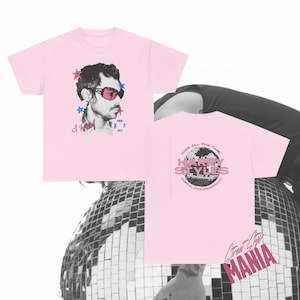 May include: A light pink short-sleeve t-shirt featuring a graphic of a person wearing pink sunglasses on the front. The back displays a disco ball graphic with the text HARRY STYLES, KISS ALL THE TIME, and DISCO, OCCASIONALLY printed in black.