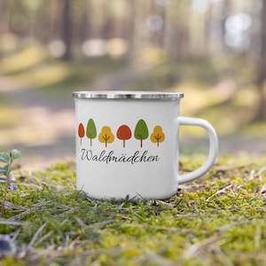 Könnte beinhalten: Ein weißer Emaillebecher mit silbernem Rand und Henkel, verziert mit einer Reihe kleiner, stilisierter Bäume in Grün-, Orange- und Gelbtönen, wobei der schwarze kursive Schriftzug "Waldmädchen" direkt unter der Baumgrafik auf der Vorderseite steht.