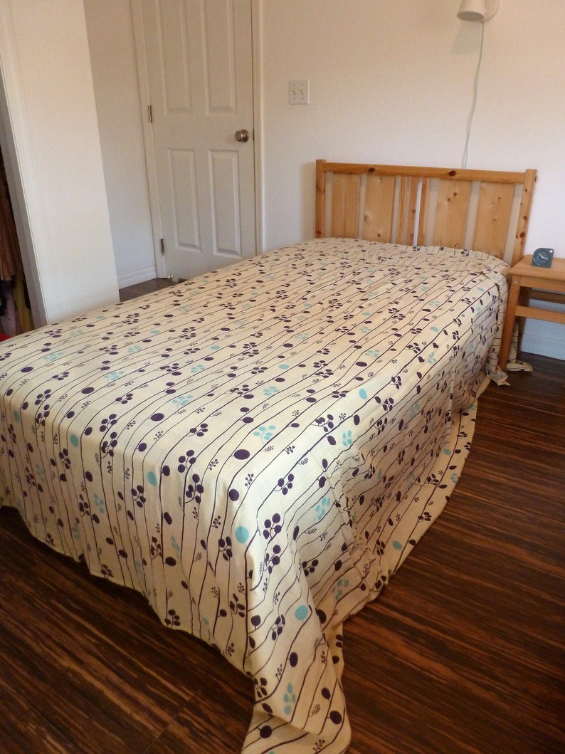 Puede incluir: Una cama blanca con una cabecera de madera y un edred&oacute;n estampado con un dise&ntilde;o floral azul claro y morado. La cama est&aacute; en una habitaci&oacute;n con suelo de madera y una puerta blanca.