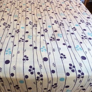 Puede incluir: Un cubrecama de color crema con un patr&oacute;n repetitivo de l&iacute;neas verticales, peque&ntilde;os c&iacute;rculos azules y formas de flores moradas.