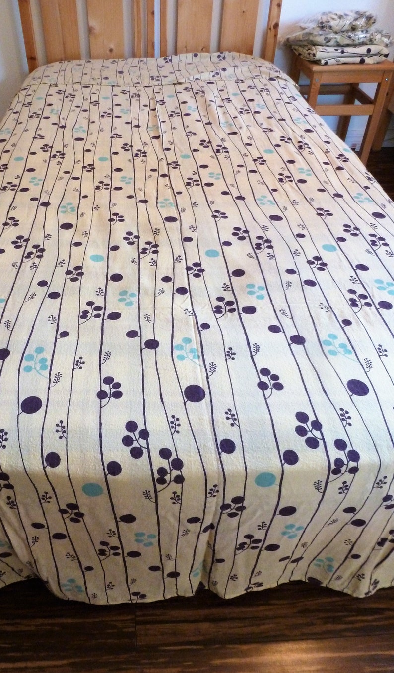Puede incluir: Un cubrecama de color crema con un patr&oacute;n repetitivo de l&iacute;neas verticales, peque&ntilde;os c&iacute;rculos azules y formas de flores moradas.