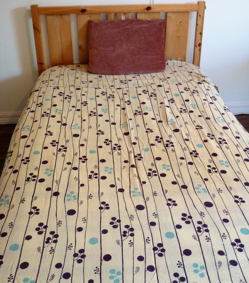 Puede incluir: Un marco de cama de madera con una cabecera y un piecero de color marr&oacute;n claro. La cama est&aacute; hecha con una colcha estampada en blanco y azul con una almohada marr&oacute;n oscuro en la parte superior.